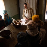 Lapsed ümber laua, laua otsas istub taluperenaine Jaanika Kuusk, puhta käteräti all on värske leib.